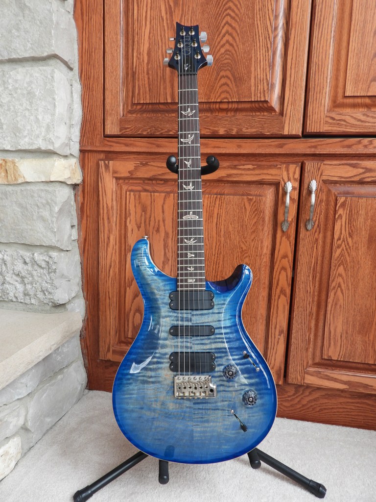 Blue electric guitar, with horizontal stripes, standing vertically on a guitar stand on the floor. 