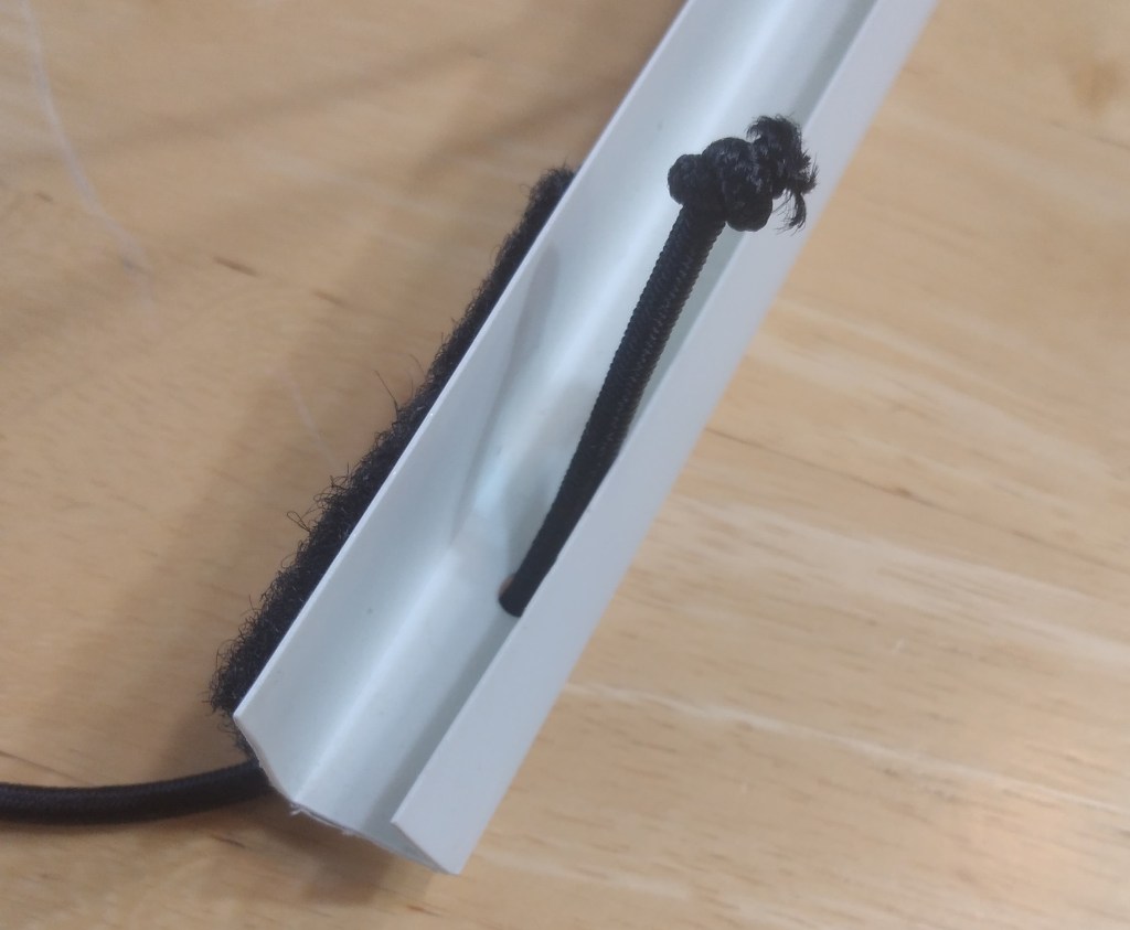 The white J-channel, which had holes drilled in it so that the black cord can pass through and be tied off with a knot.