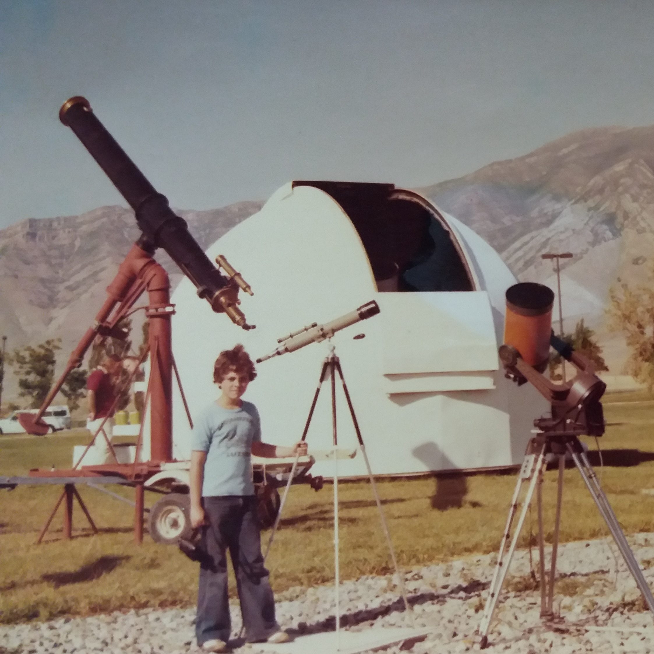Large and small astronomical telescopes, standing near a white observatory dome. One adult to the right, on the left, a child with a telescope taller than him, frizzy brown hair, blue t-shirt, bell-bottom jeans and sneakers. Late afternoon sun, mountains in the background.